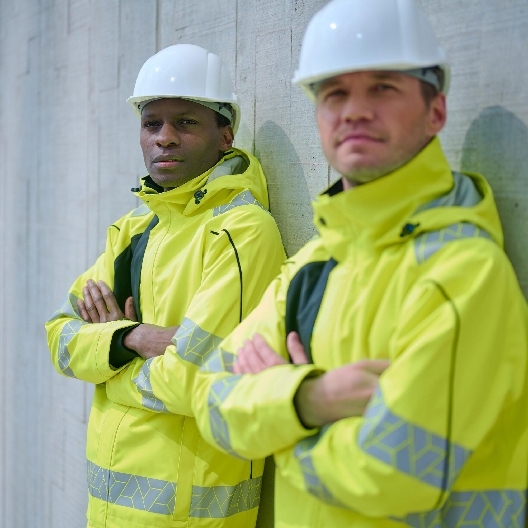 Two construction workers wearing bright yellow high-visibility jackets with reflective strips and white safety helmets stand against a concrete wall, showcasing workplace safety clothing suitable for construction and industrial environments.