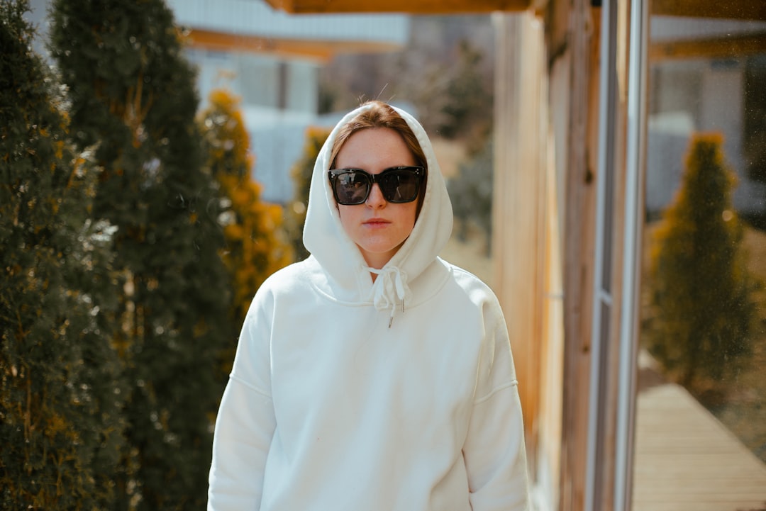 A person wearing a white hoodie with the hood up and black sunglasses, walking outdoors along a modern walkway with greenery and buildings in the background under warm daylight.