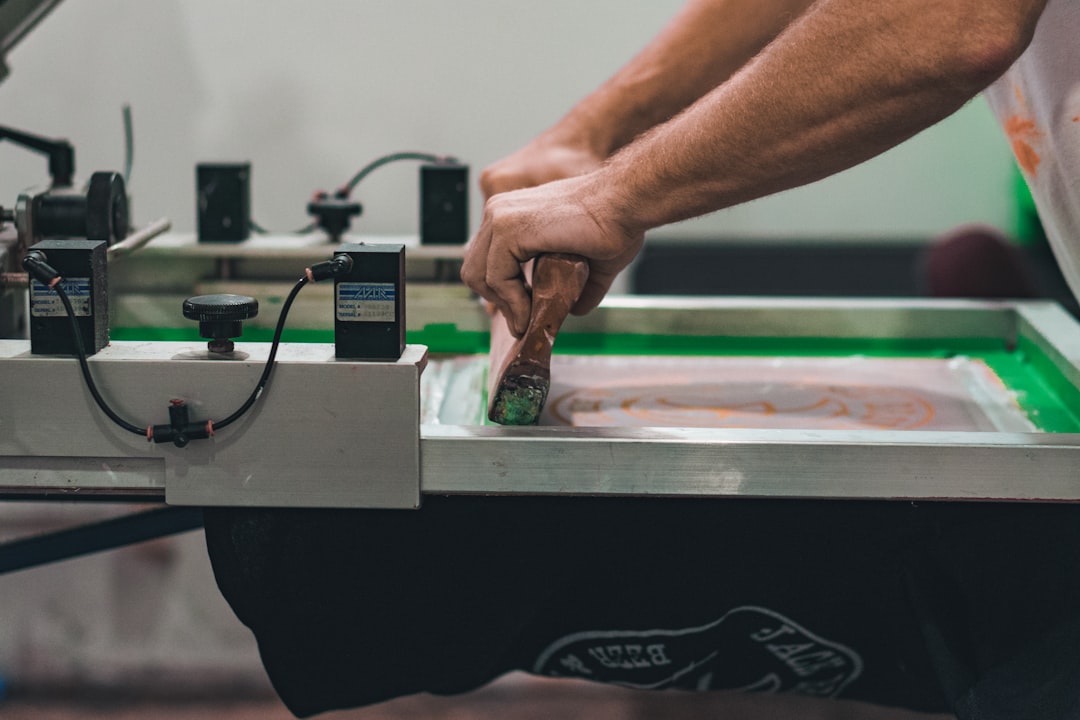 Close-up of hands using a squeegee to apply ink during a screen printing process on a mesh screen, showing industrial printing equipment and textile printing workflow